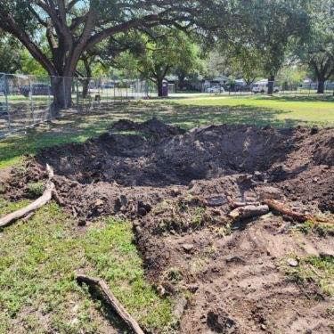 Large dirt hole in a grassy park, surrounded by upturned soil and scattered debris. Trees and a fence are in the background.