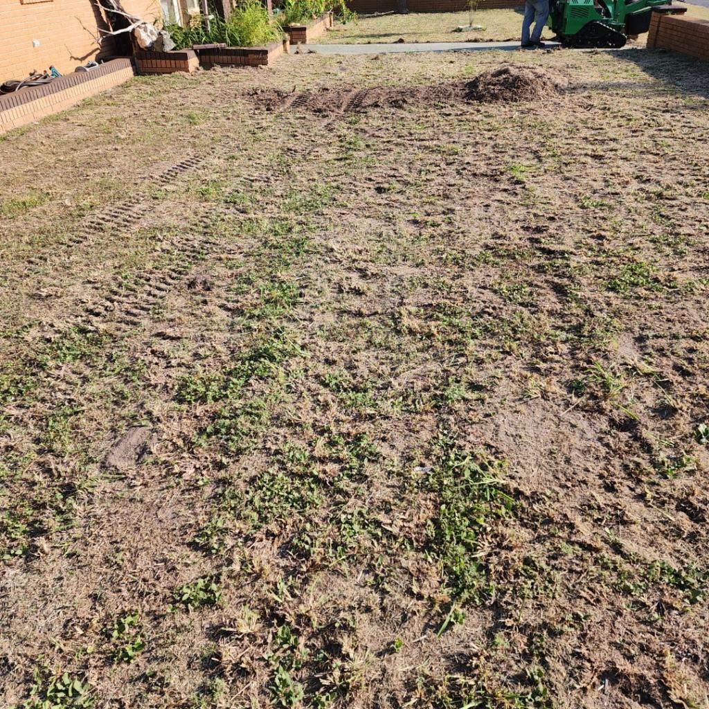 A bare, patchy lawn with small weeds, dirt tracks, and a machine in the background, possibly aerating the soil.