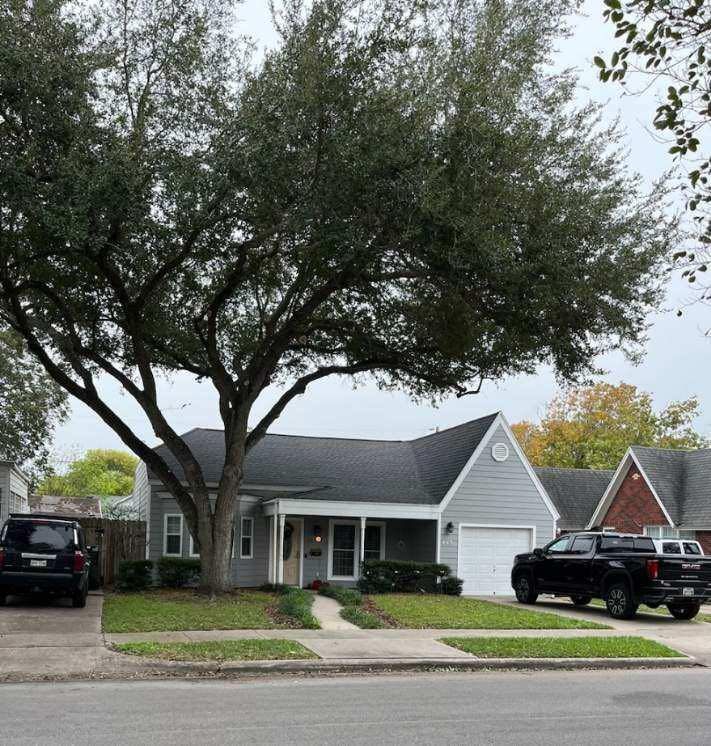 Small gray house with a large tree in front, cars in driveway, and a cloudy sky.