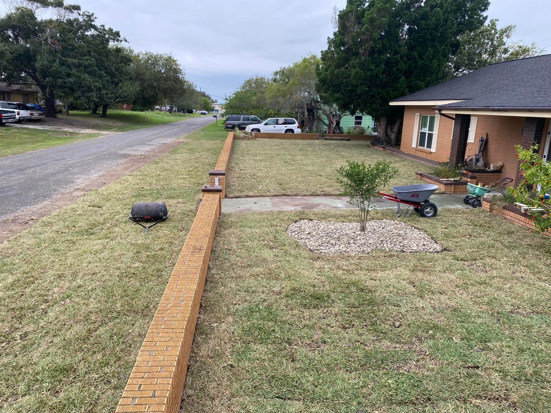 A residential street with a brick retaining wall, grass, a small tree, and a lawnmower.