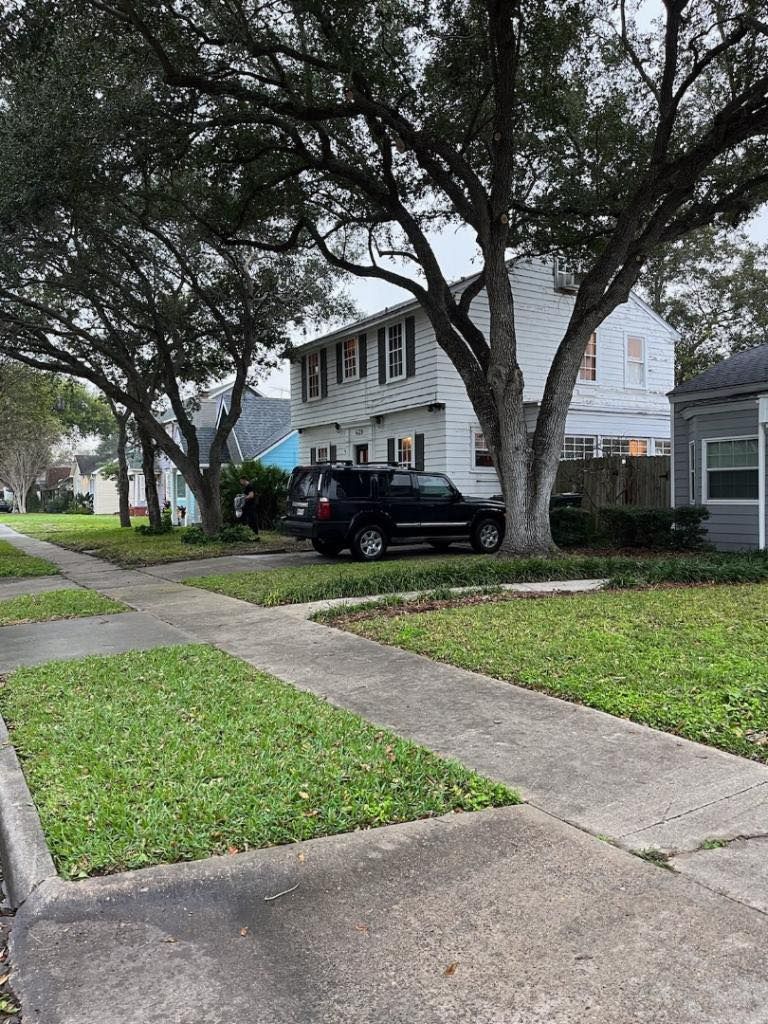 Street scene: white house with black SUV parked in front, lush green lawns, sidewalk, large trees.