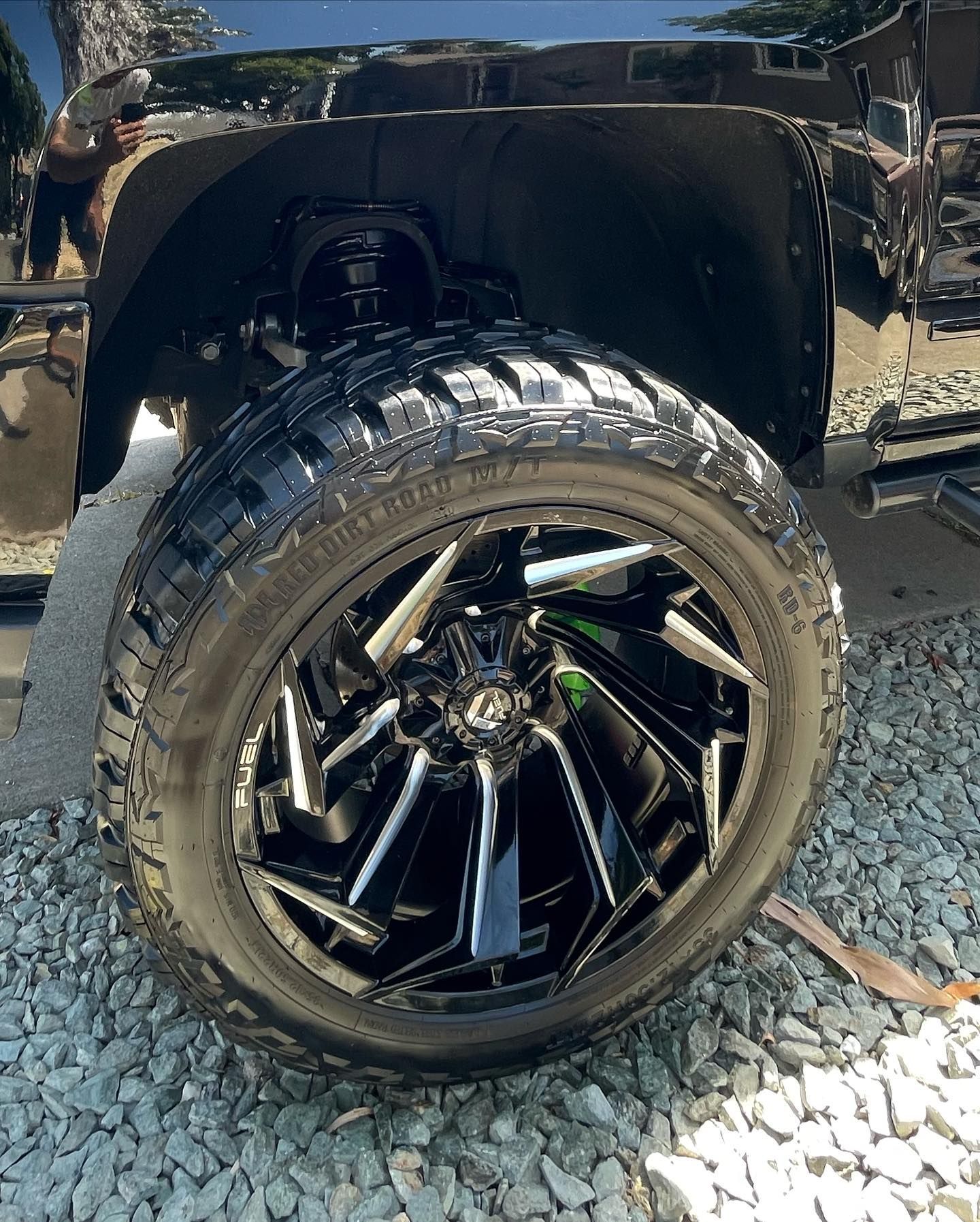 A black truck with black wheels and tires is parked on gravel.