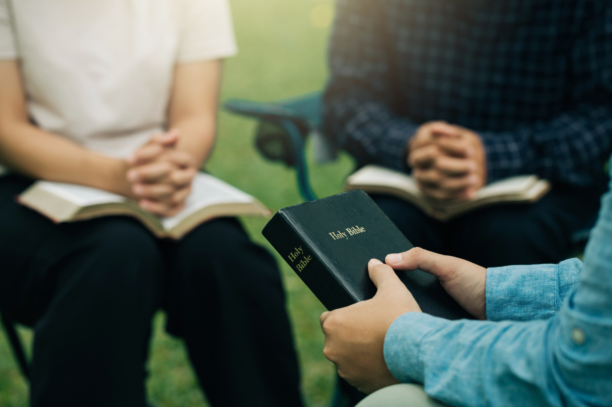 Three people sitting and reading from books, one holding a black Bible.