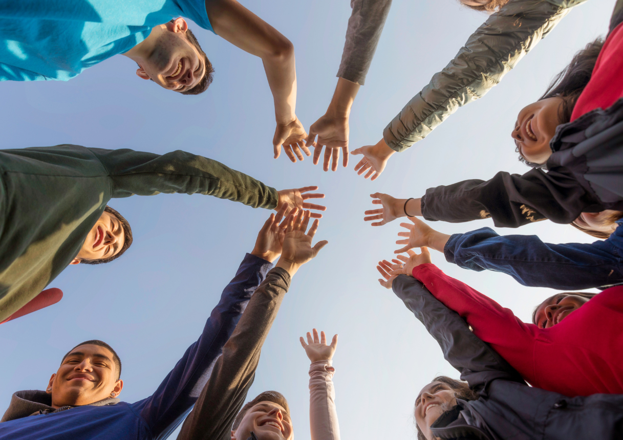 Group of people with arms raised, hands touching in a circle against a blue sky.