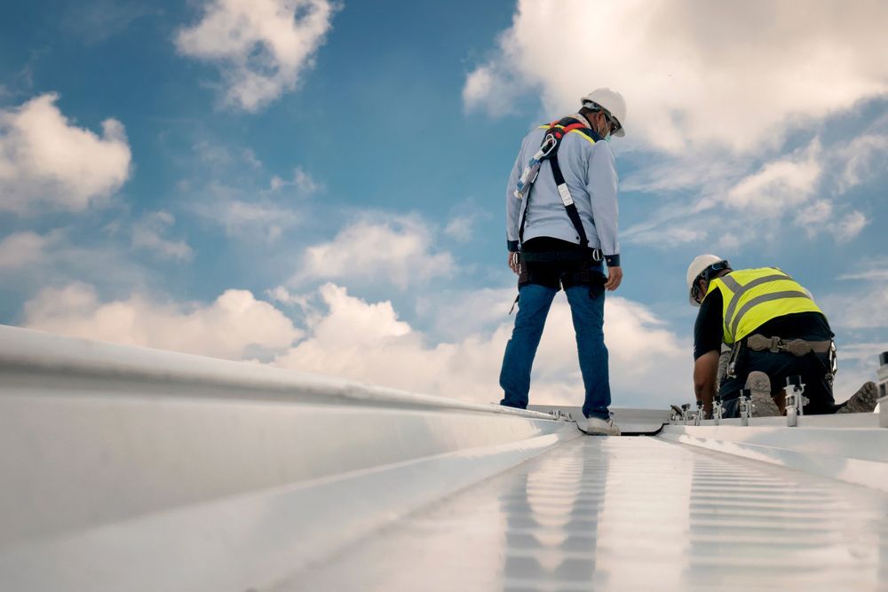 Two construction workers wearing safety gear standing and kneeling on a metal roof.