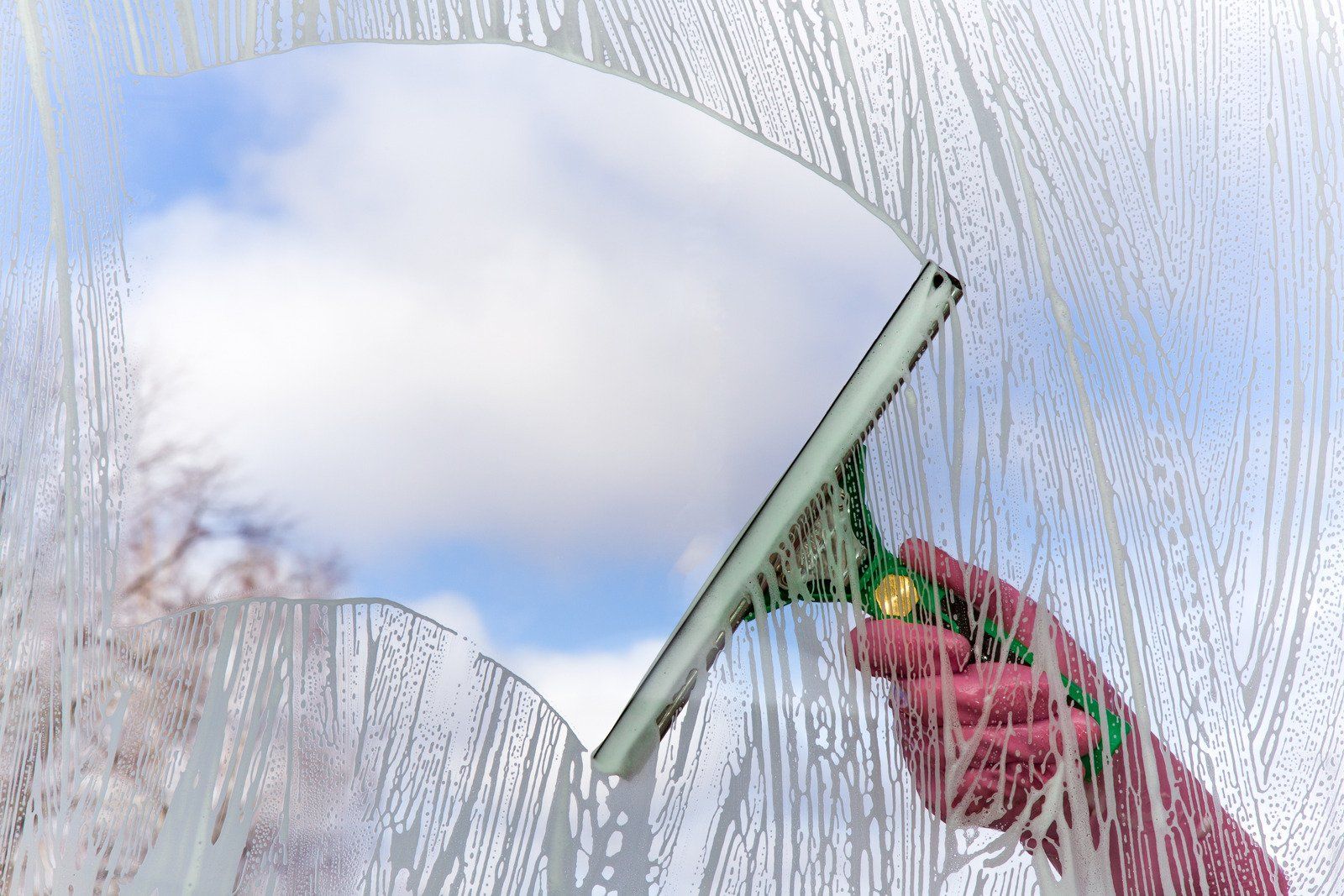 A person wearing pink gloves cleans a window with a squeegee, revealing a blue sky with clouds.