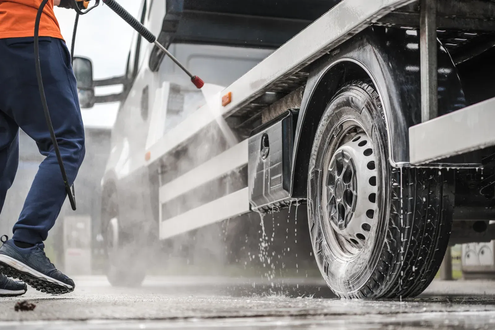 Person washing a white commercial van's tire with a pressure washer.