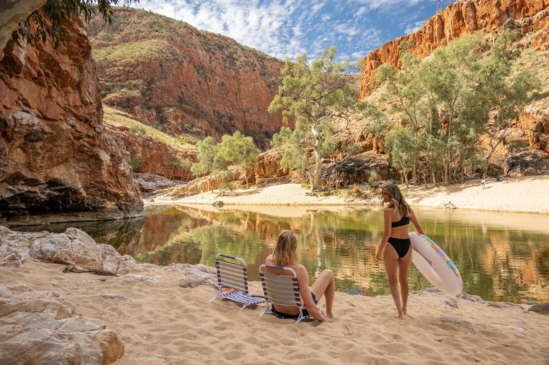 Two women in bikinis are sitting on a beach next to a river.