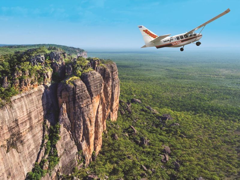 A small plane is flying over a cliff with trees on it