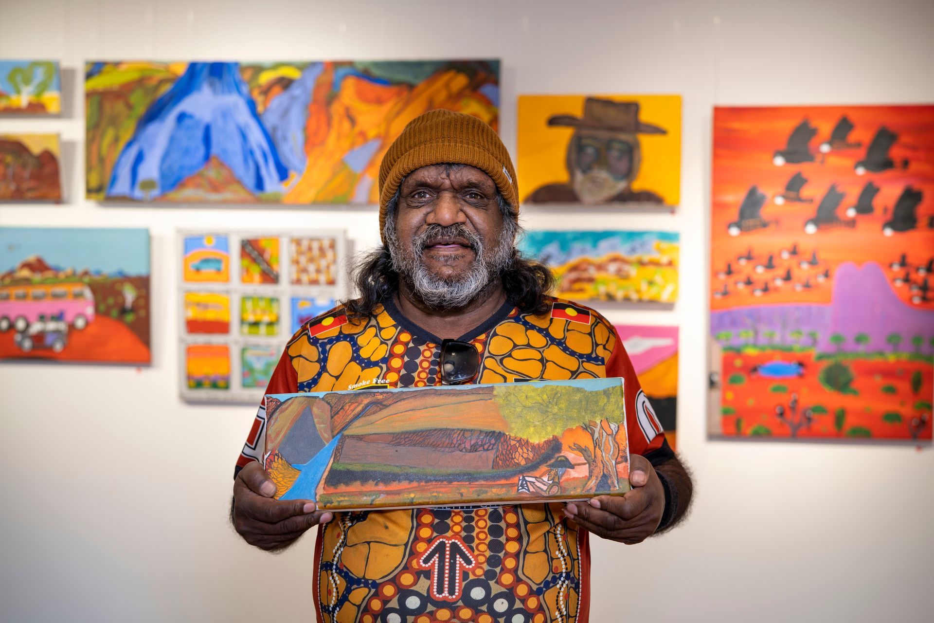 A man is holding a painting in front of a wall of paintings.