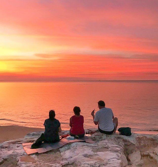 Three people are sitting on a rock watching the sunset over the ocean.