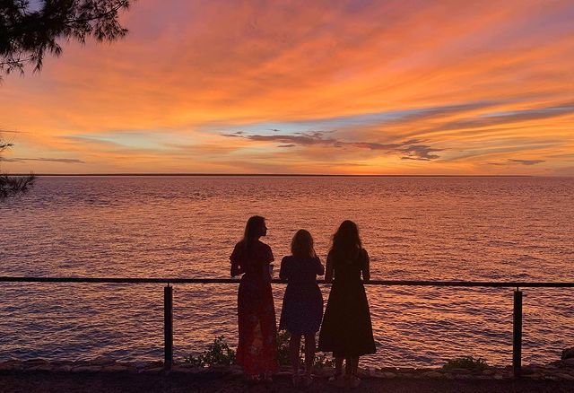 Three women are standing on a balcony overlooking the ocean at sunset.