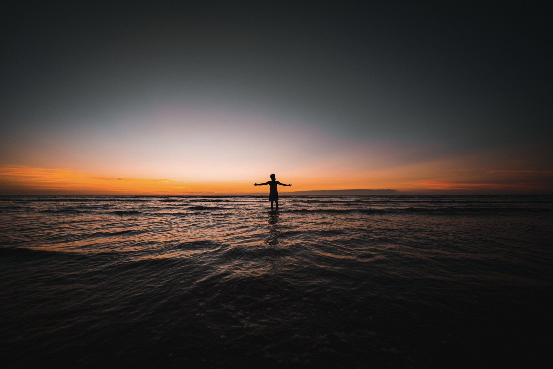 A person is standing in the ocean at sunset with their arms outstretched.
