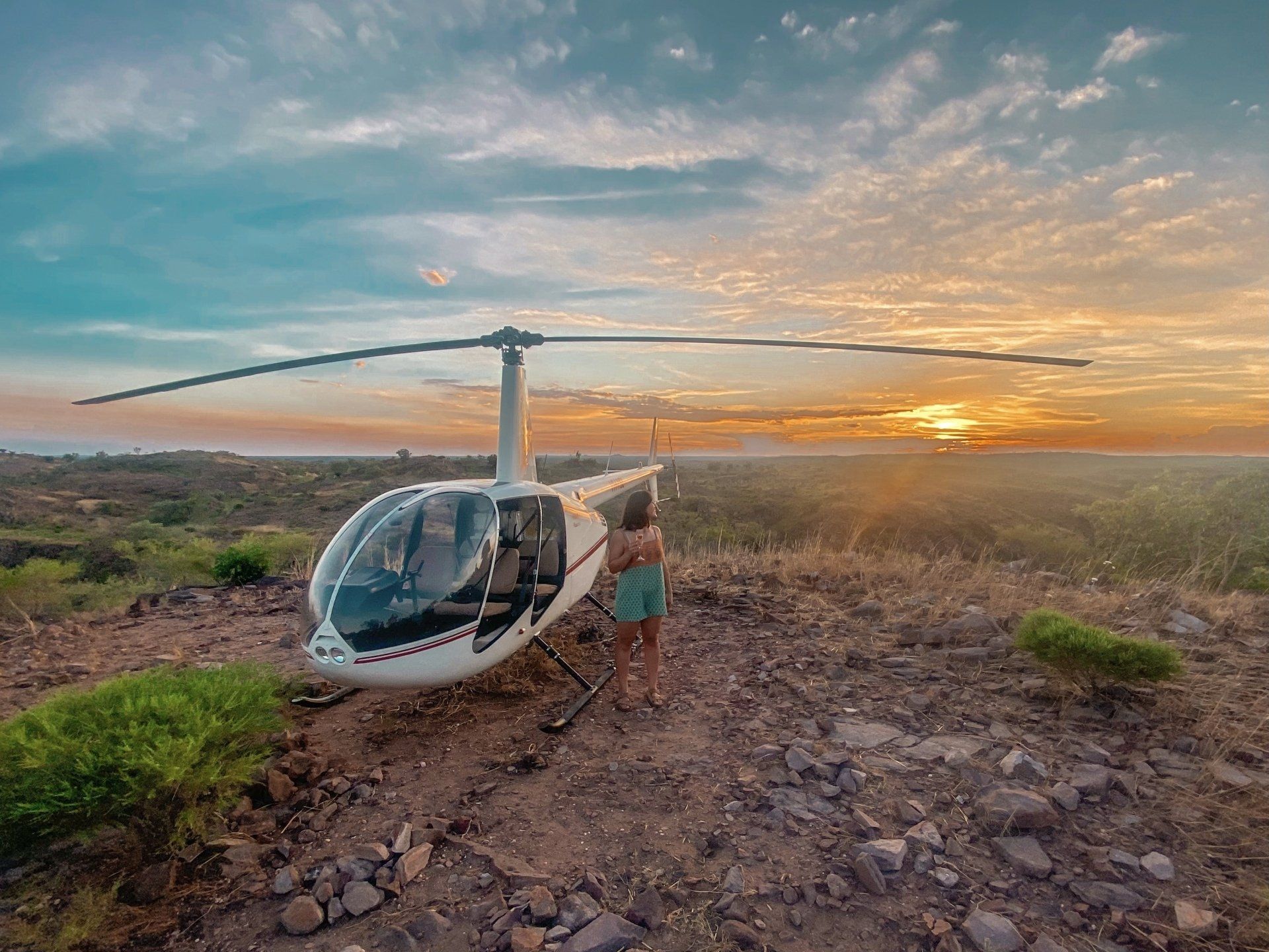 A woman is standing next to a helicopter on top of a hill at sunset.