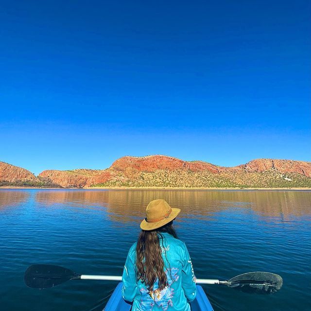 A woman is sitting in a kayak on a lake with mountains in the background.