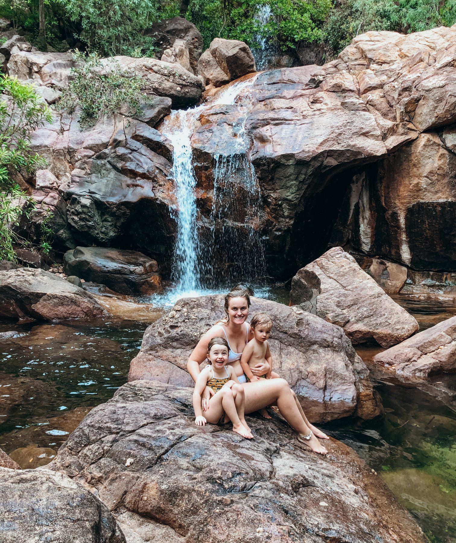 A woman and two children are sitting on a rock in front of a waterfall.