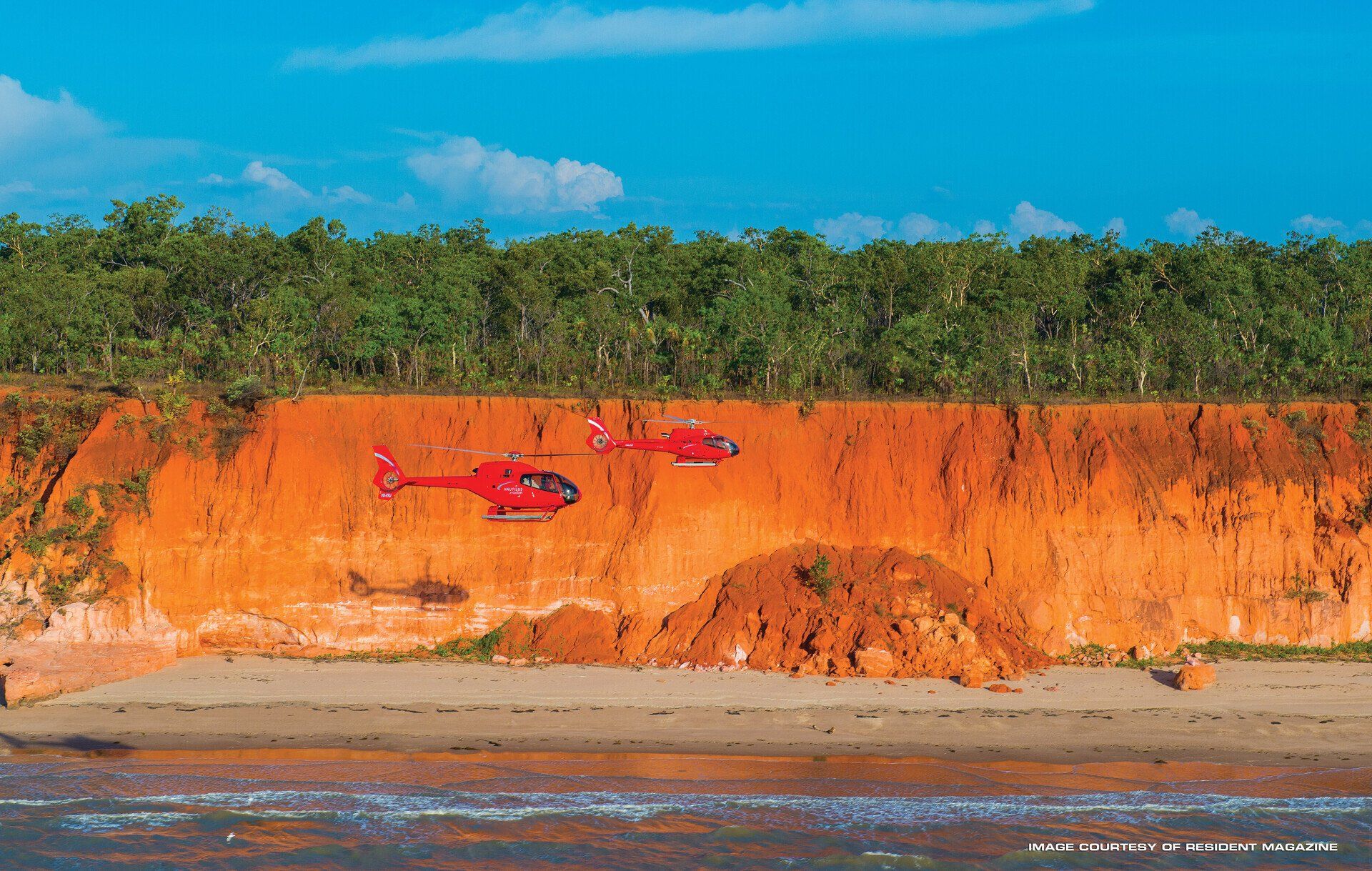 Two red helicopters are flying over a sandy beach.