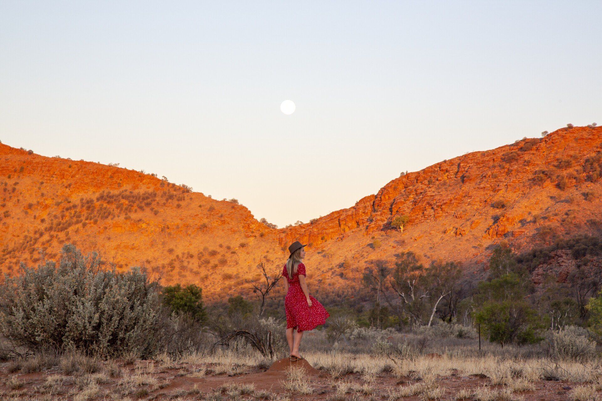 A woman in a red dress is standing in the desert looking at the moon.