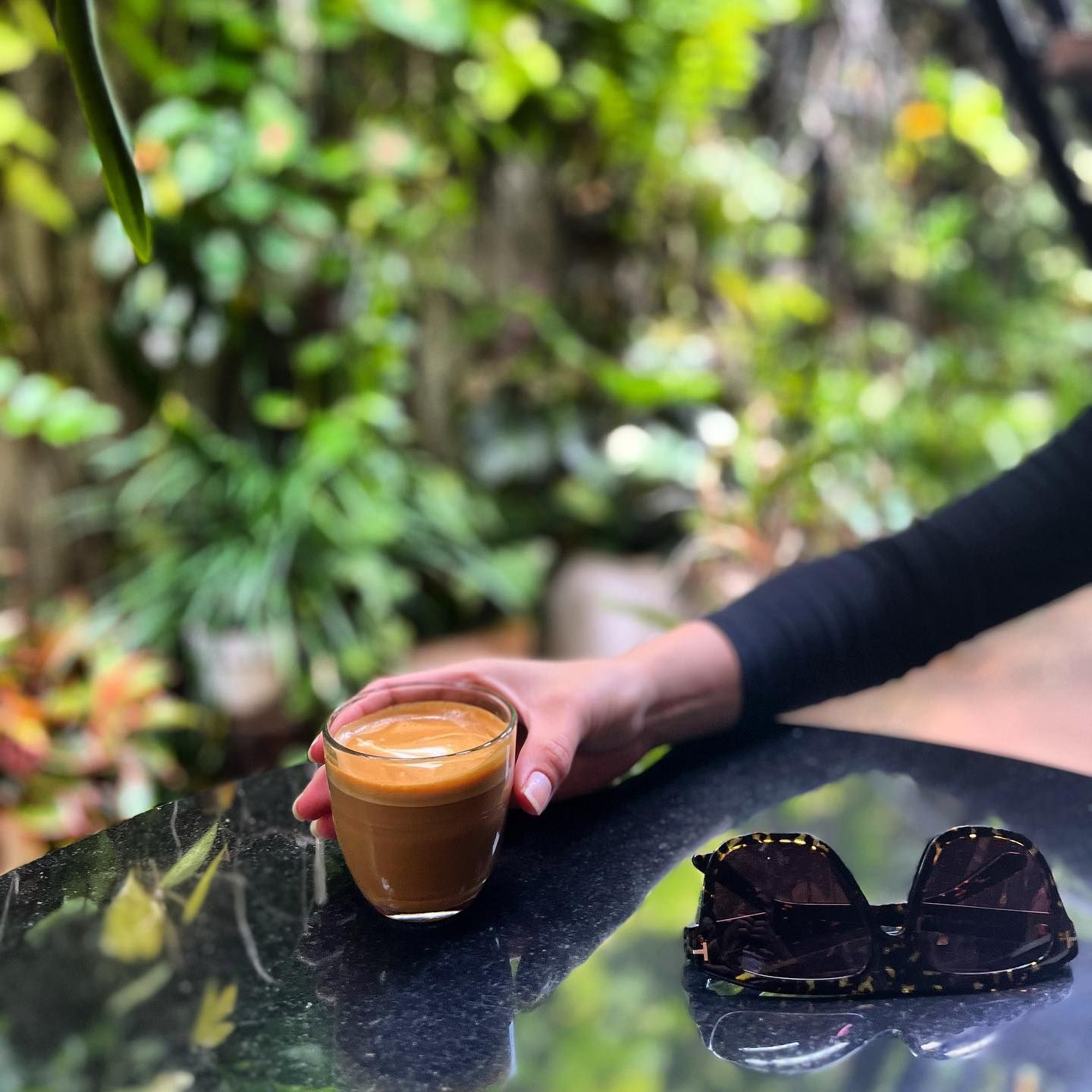 A person is holding a cup of coffee on a table next to sunglasses.