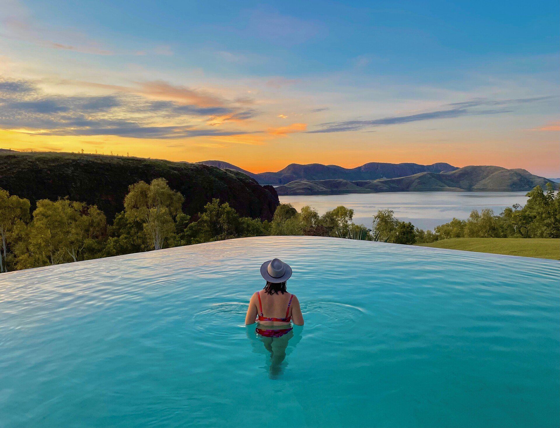 A woman is swimming in an infinity pool at sunset.