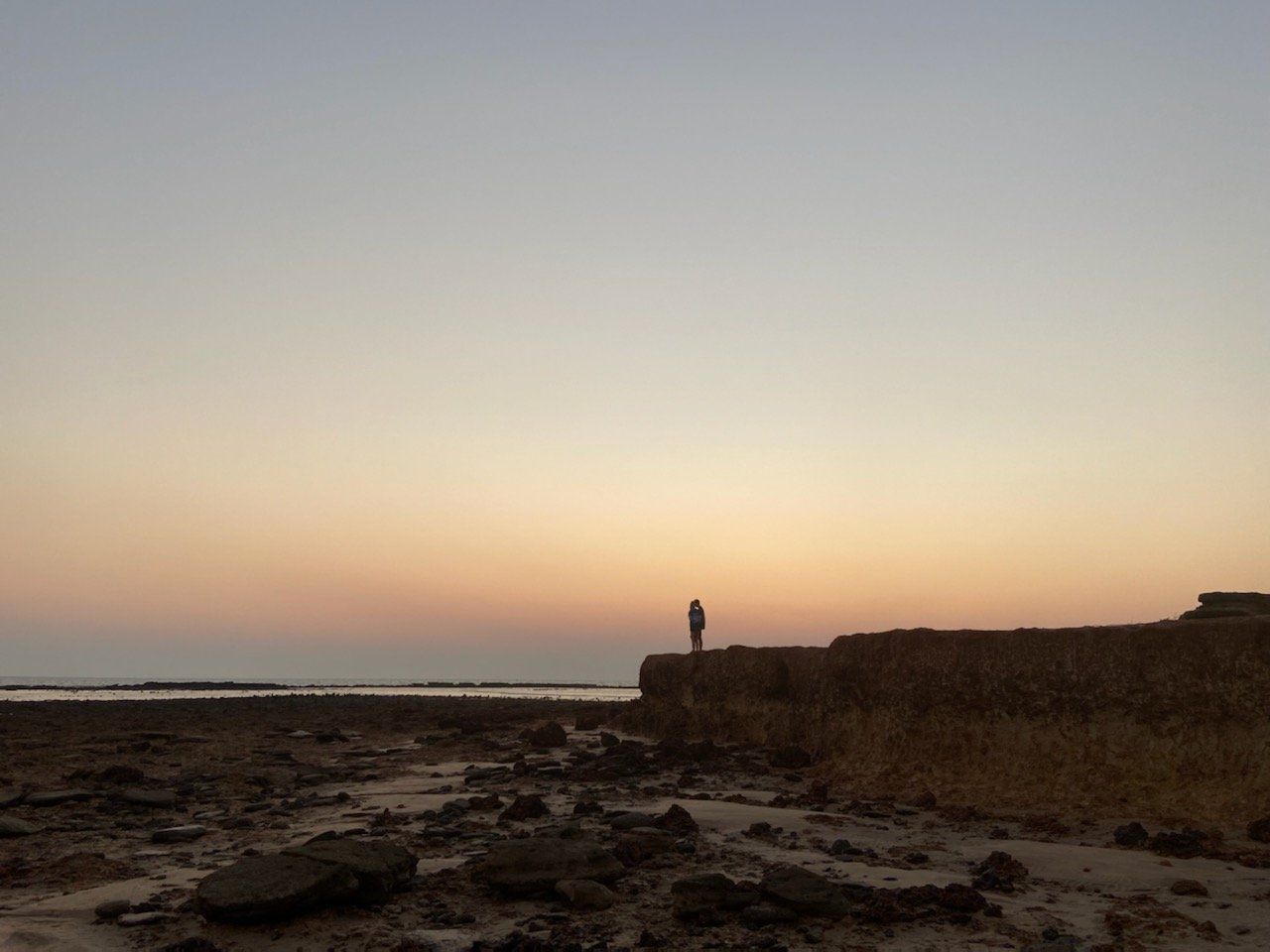 A person is standing on a rocky beach at sunset.