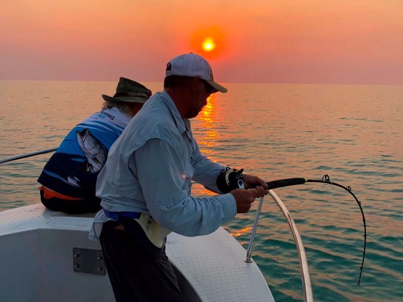 Two men are fishing on a boat with the sun setting in the background