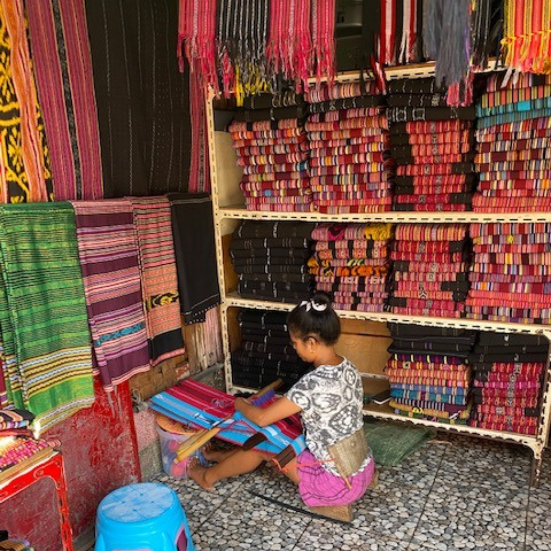 A woman is working on a loom in a store filled with lots of colorful fabrics.
