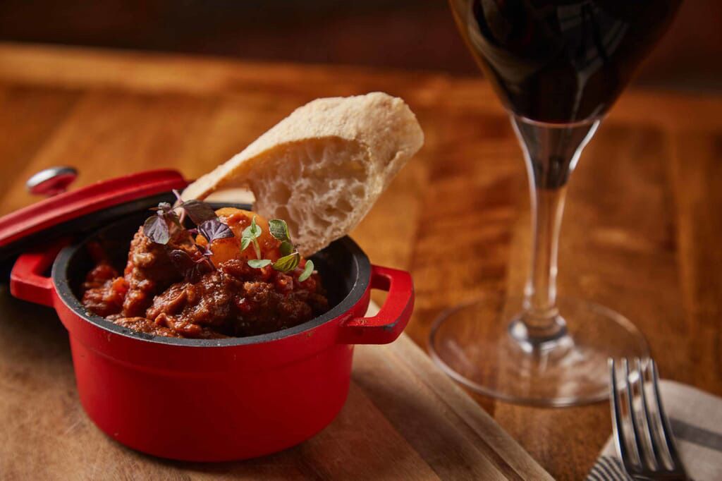 a red pot filled with meat and bread next to a glass of wine