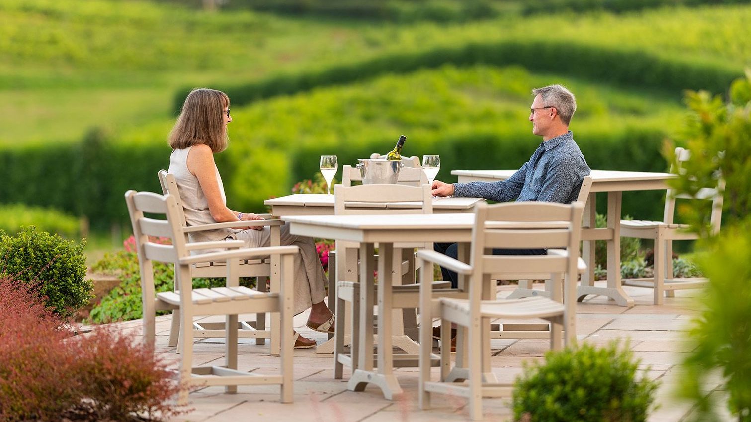 A couple sits at a table outdoors in a vineyard, enjoying a meal. Green foliage and rolling hills are in the background.