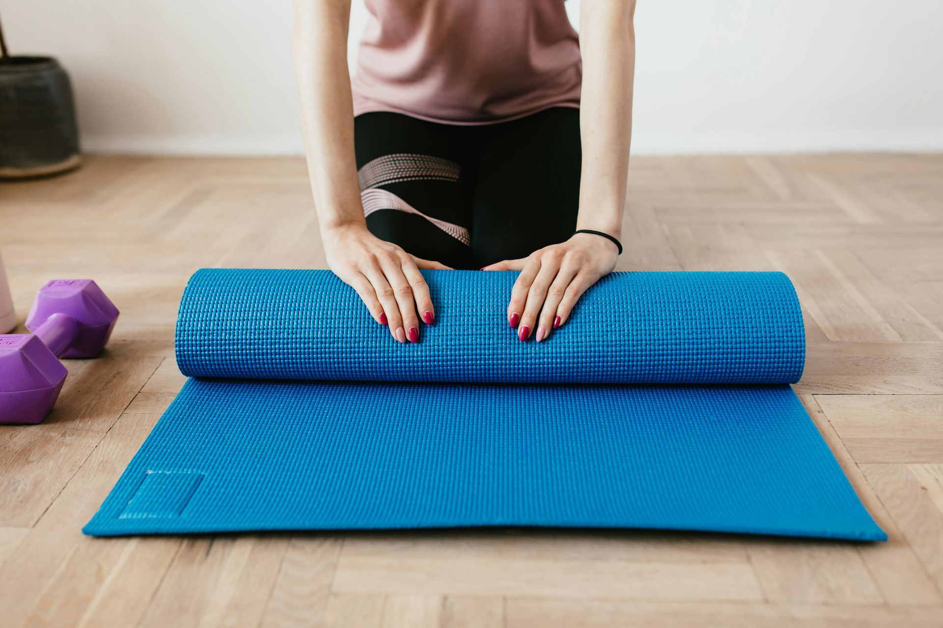 Person rolling out a blue yoga mat on a wooden floor, purple dumbbells in the background.