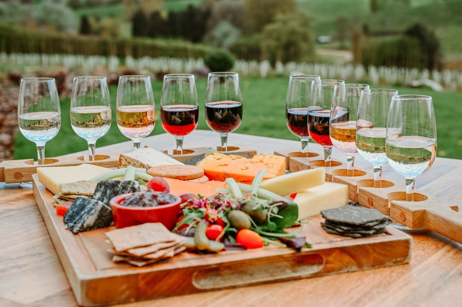 A wooden board with cheese, crackers, and appetizers, with wine glasses set around it on a table outdoors in a vineyard.
