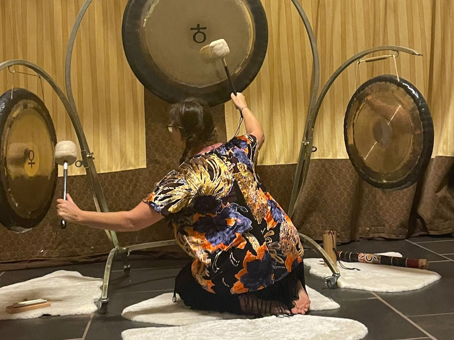 A person kneels, striking gongs with mallets. They are indoors, amidst draped fabric and decorative gongs.