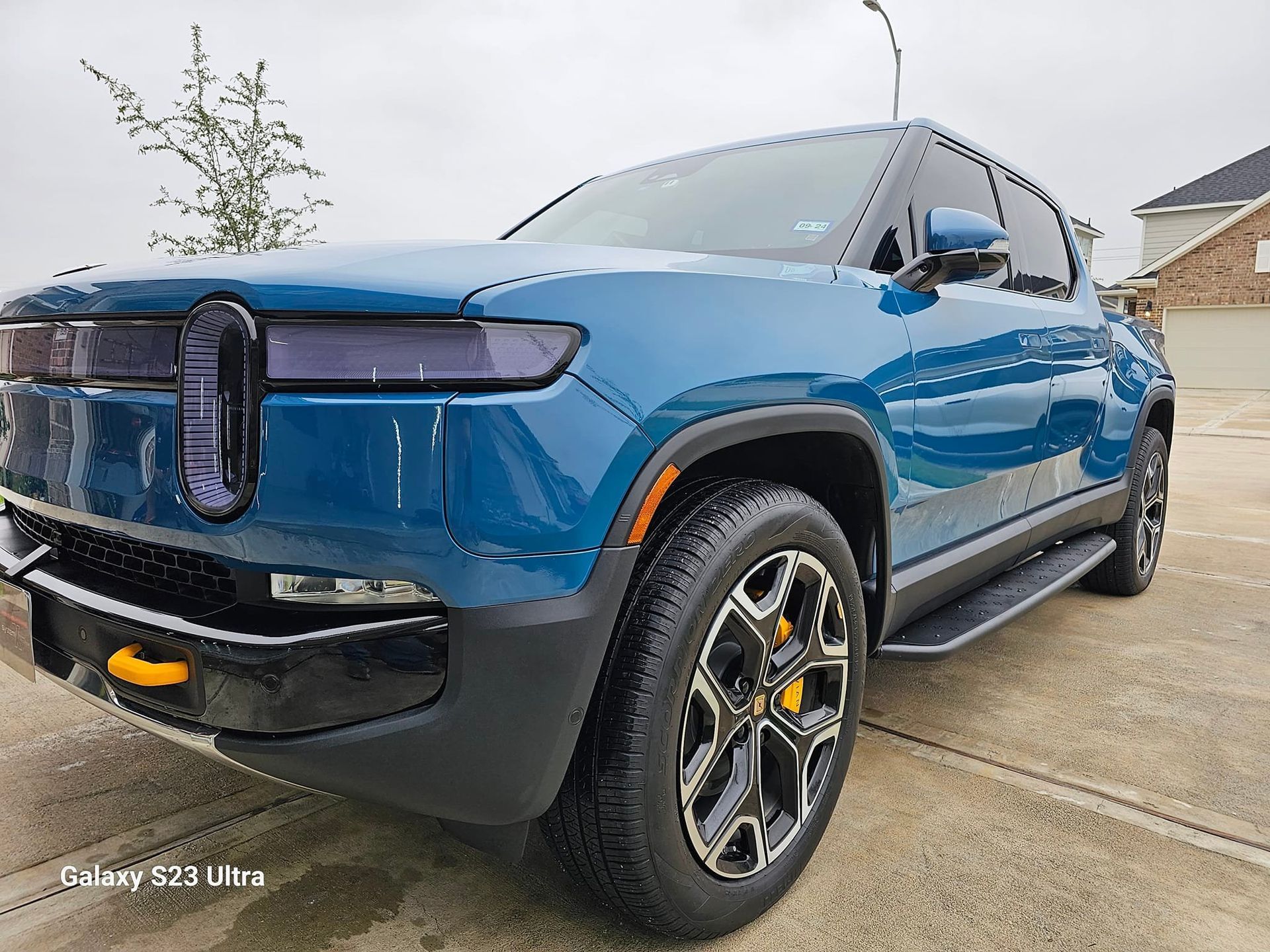 A blue Rivian R1T electric pickup truck parked on a concrete driveway with a house in the background.