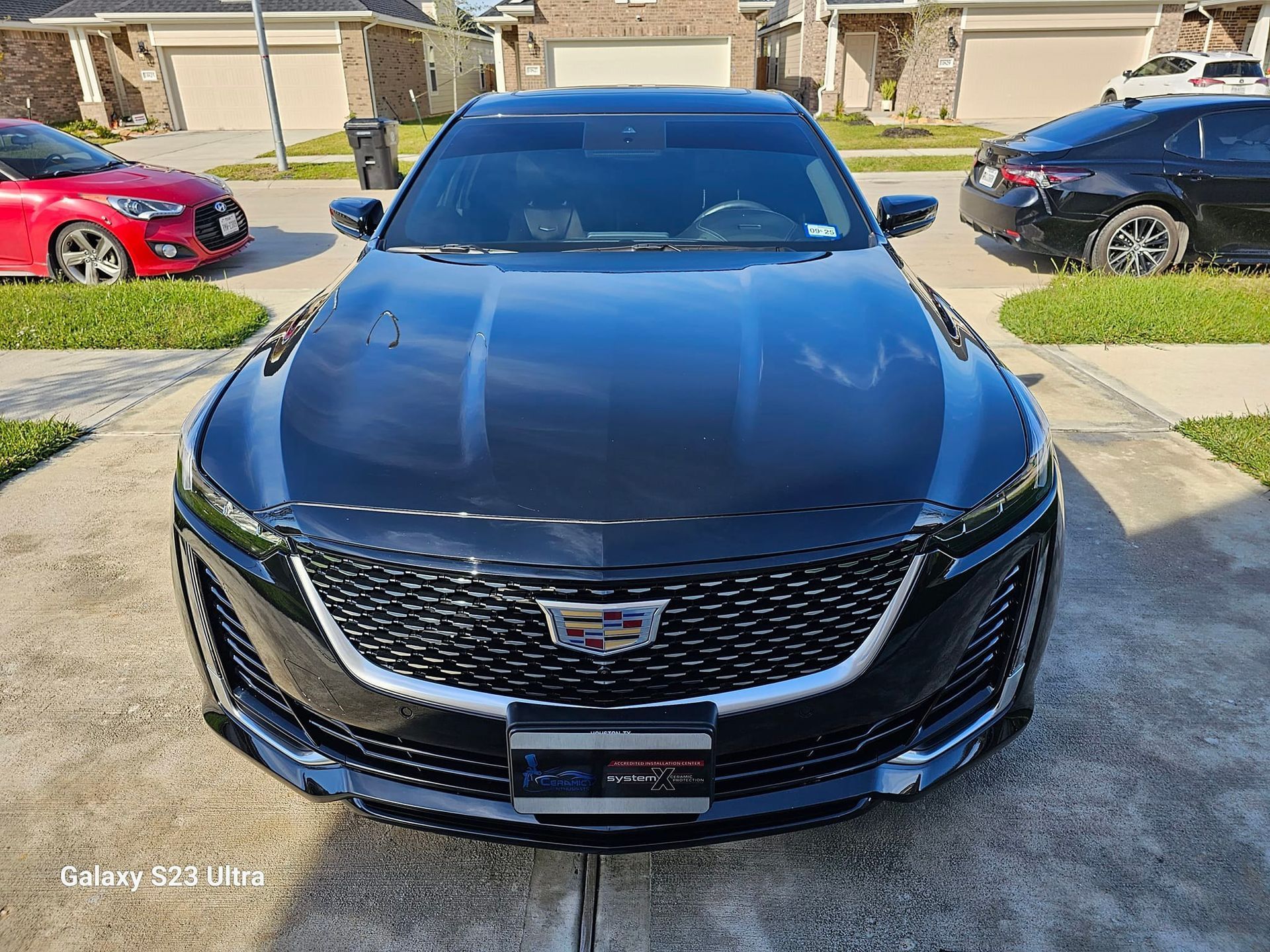 Front view of a shiny, black Cadillac sedan parked in a suburban driveway on a sunny day.