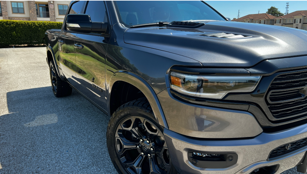 A grey Ram pickup truck parked on a paved lot on a sunny day, showcasing its front grille, headlights, and large wheels.