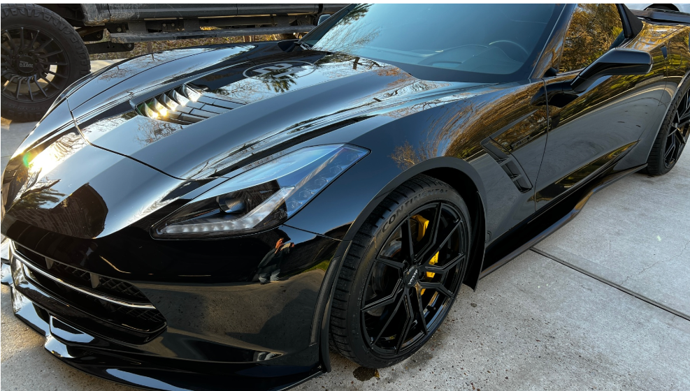 A black Chevrolet Corvette C7 sports car parked outdoors, featuring yellow brake calipers and black alloy wheels.