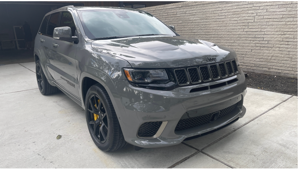 A gray Jeep Grand Cherokee Trackhawk parked on a concrete driveway in front of a light-colored brick wall.