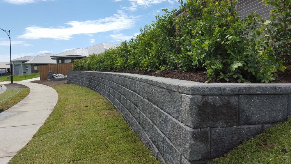 A Stone Wall Surrounds A Lush Green Lawn In Front Of A House — Green Envy Landscape & Garden Services In Cameron Park NSW
