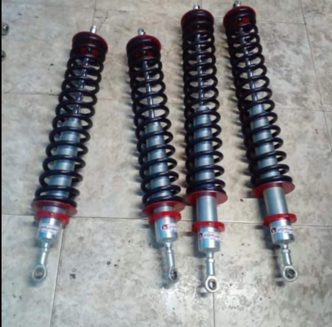 Cuatro amortiguadores tipo coilover con resortes negros, cuerpos plateados y detalles en rojo.