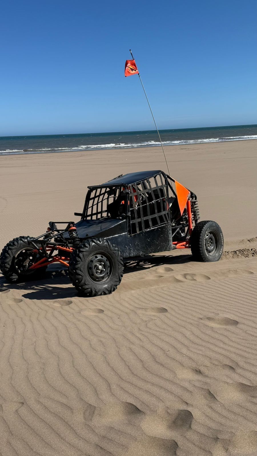 Buggy de dunas negro y naranja en una playa de arena bajo un cielo azul, con una bandera de seguridad.