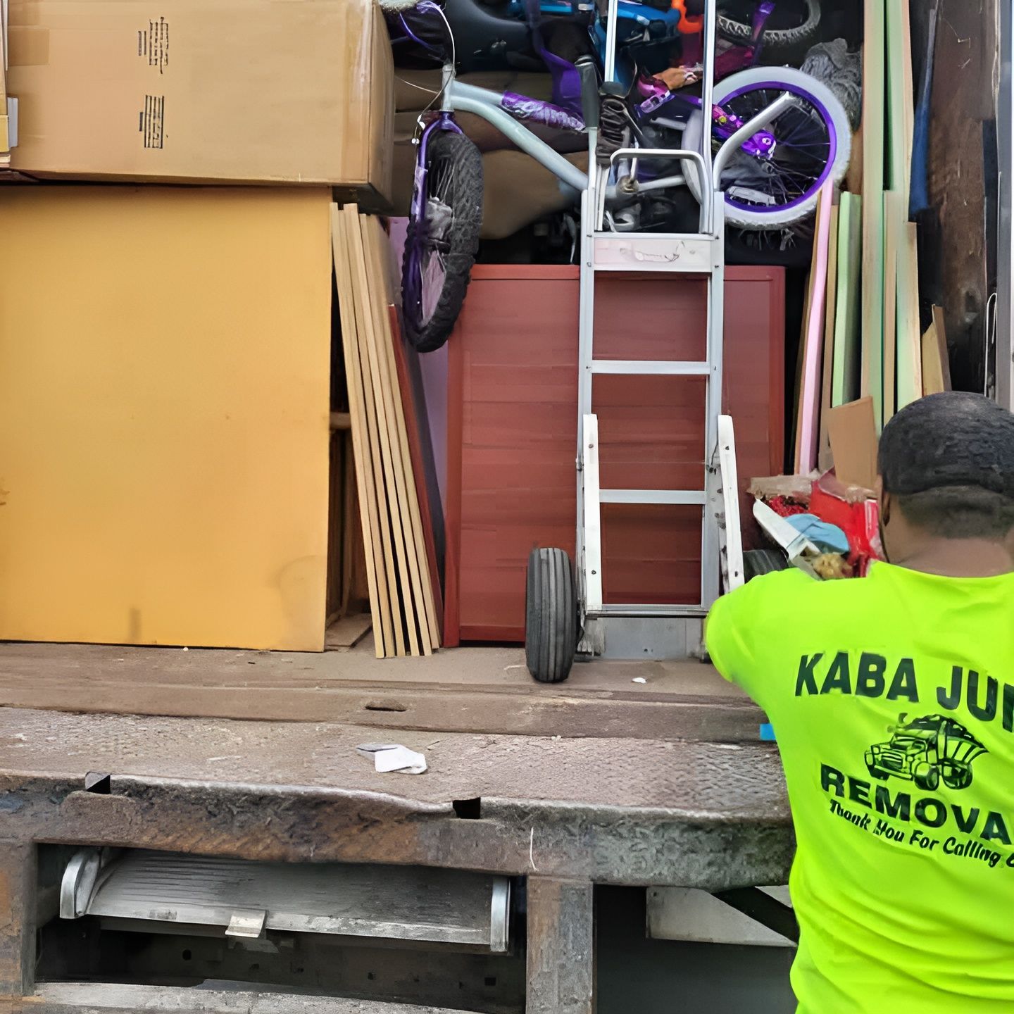 A man in a neon green kaba junk removal shirt is loading a truck with boxes and a ladder.