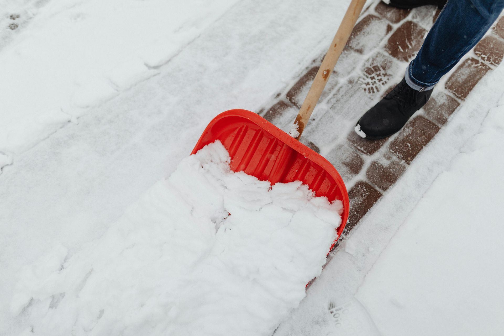 A person is shoveling snow on a sidewalk with a red shovel.