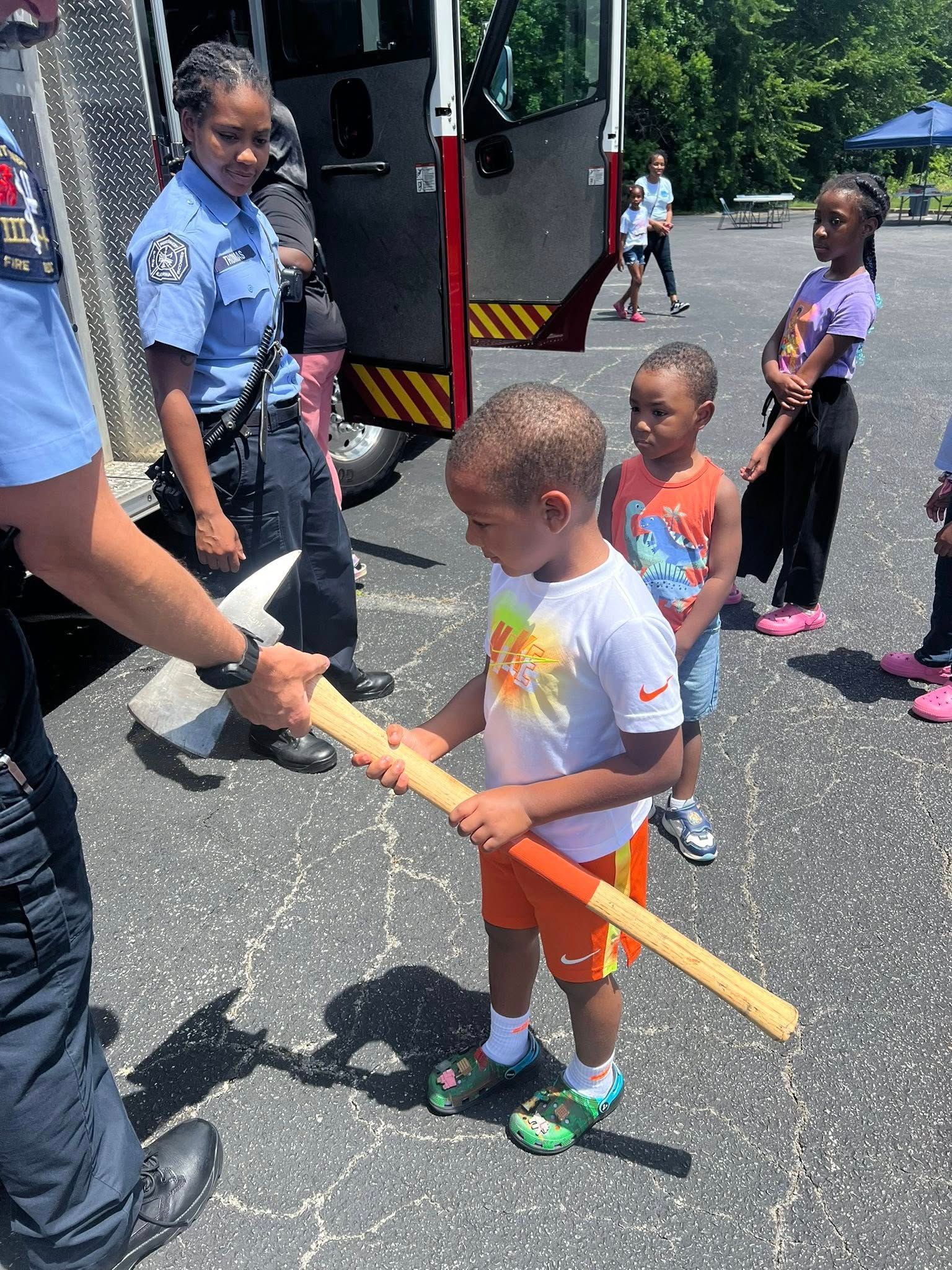 A little boy is holding a wooden stick in front of a fire truck.