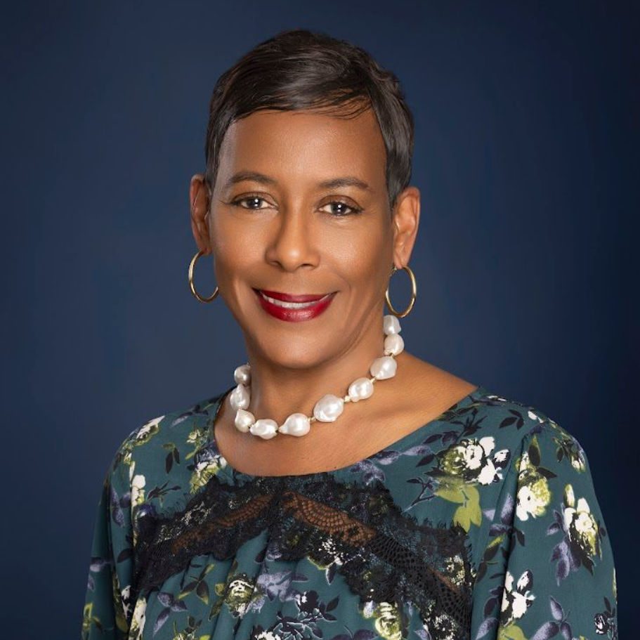 Woman with short dark hair, wearing a floral top and pearl necklace, smiling on a blue background.