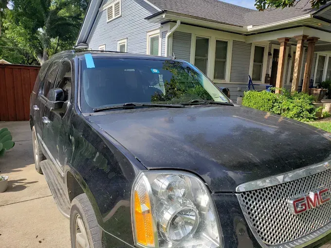 Black GMC Yukon SUV parked in front of a blue house with trees in the background on a sunny day.
