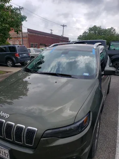 Green Jeep Cherokee parked, showing front, under cloudy sky. Buildings in background.