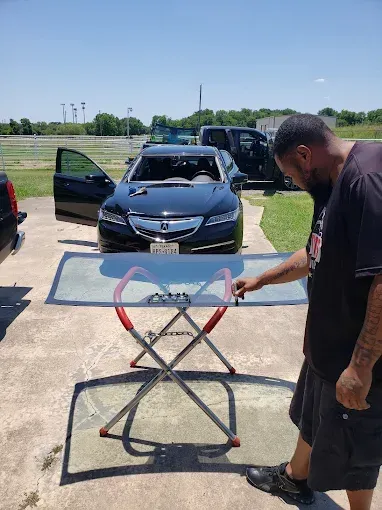 Man points at a car windshield on a folding stand, near a black car with an open door outdoors on a sunny day.