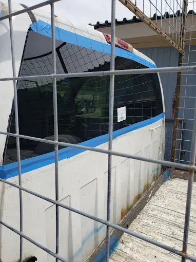 White vehicle rear side behind a wire fence, blue tape on top, windows and a glimpse of interior.