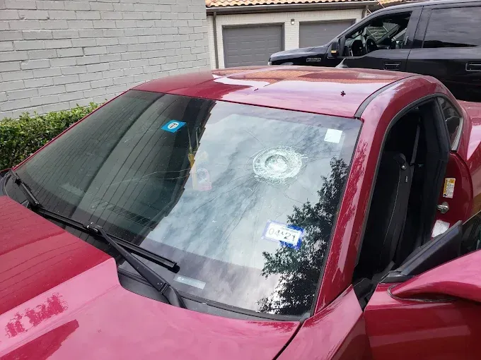 Red car windshield with a large hole, likely from impact, in front of a house.