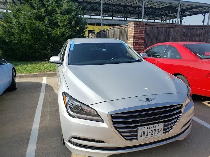 Silver Genesis sedan parked in a lot, with a red car and green foliage nearby.